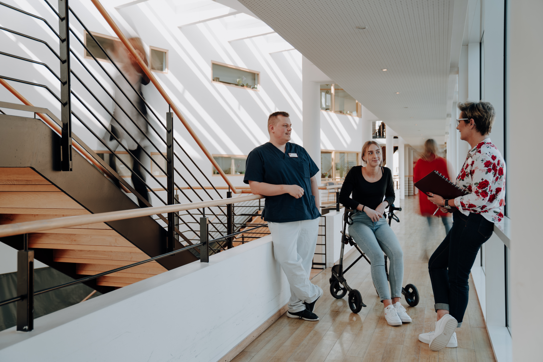 Ein Mann, der an einem Geländer lehnt, eine Frau, die auf einem Rollator sitzt, und eine Frau mit einer Mappe in der Hand stehe in einem Foyer.