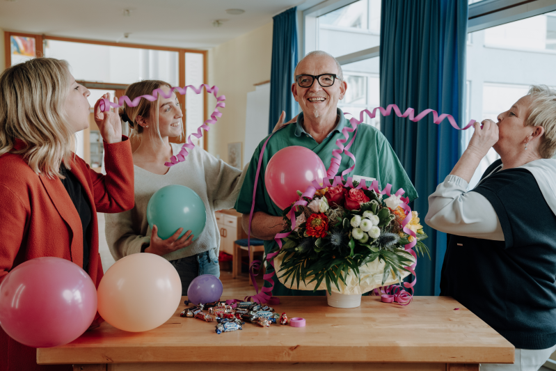3 Frauen, die Luftschlangen pusten und um einen älteren Mann stehen, der vor einem Tisch mit einem Blumenstraß und Luftballons steht