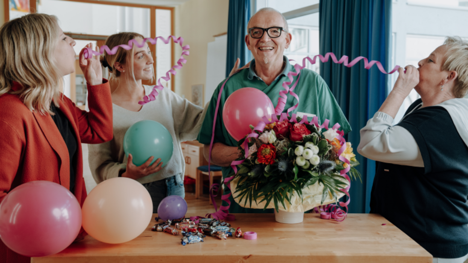 3 Frauen, die Luftschlangen pusten und um einen älteren Mann stehen, der vor einem Tisch mit einem Blumenstraß und Luftballons steht