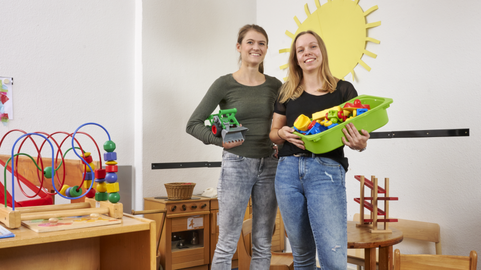 Zwei Frauen stehen im Spielbereich einer Kita und lächeln in die Kamera