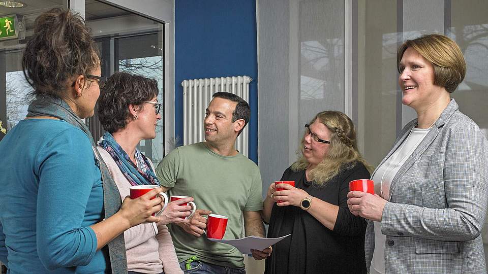 Fünf Menschen stehen zusammen, halten eine Kaffeetasse in der Hand und unterhalten sich