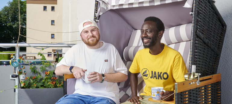 Zwei Männer sitzen mit einer Kaffeetasse in einem Strandkorb und lächeln in die Kamera