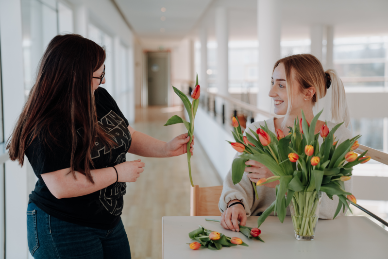Eine Frau steht hinter einer Vase mit Tulpen und gibt eine Tulpe an eine andere Frau