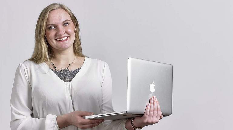 Eine Frau hält einen Laptop in der Hand und lächelt in die Kamera